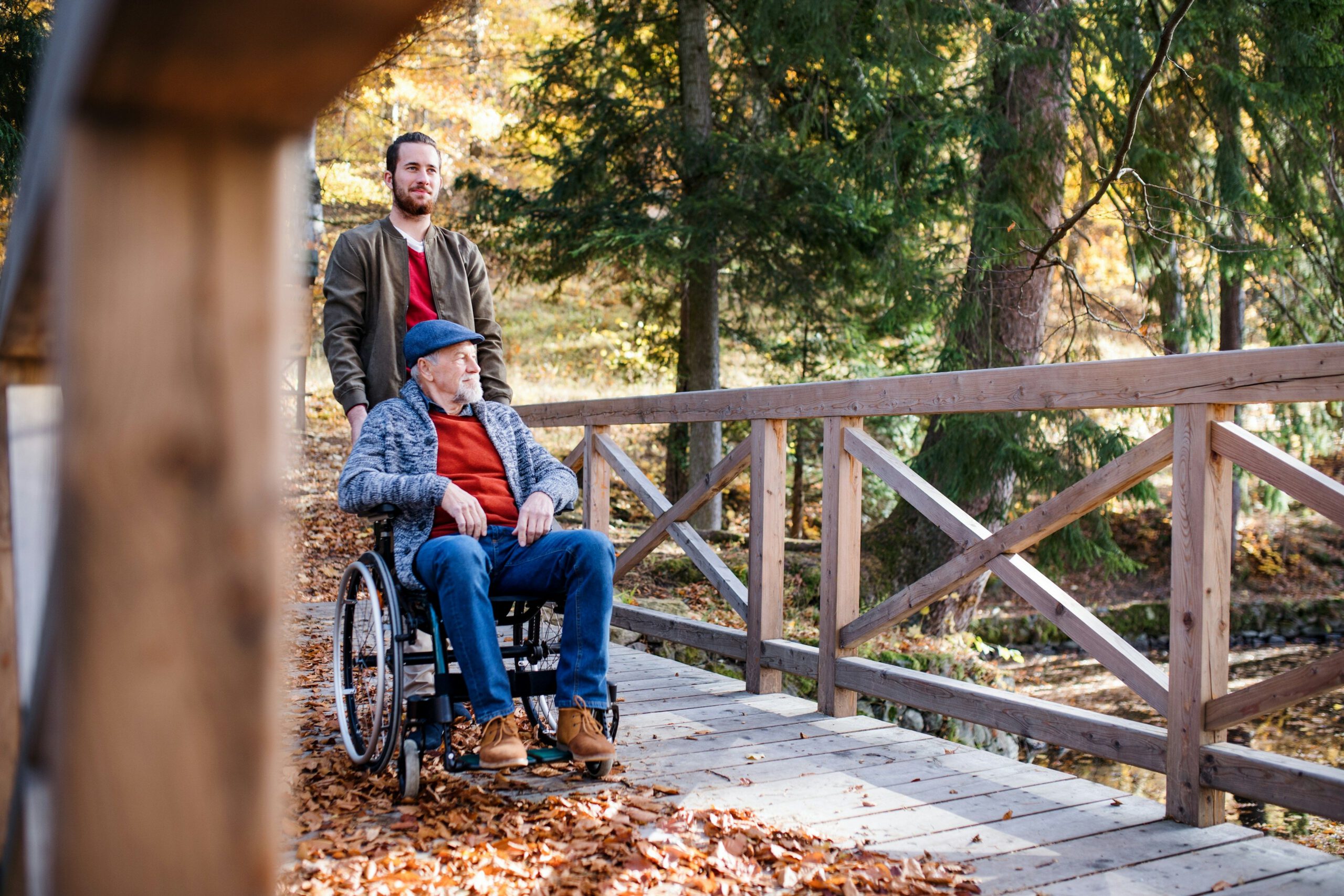 Caregiver pushing an elderly man in a wheelchair across a wooden bridge outdoors