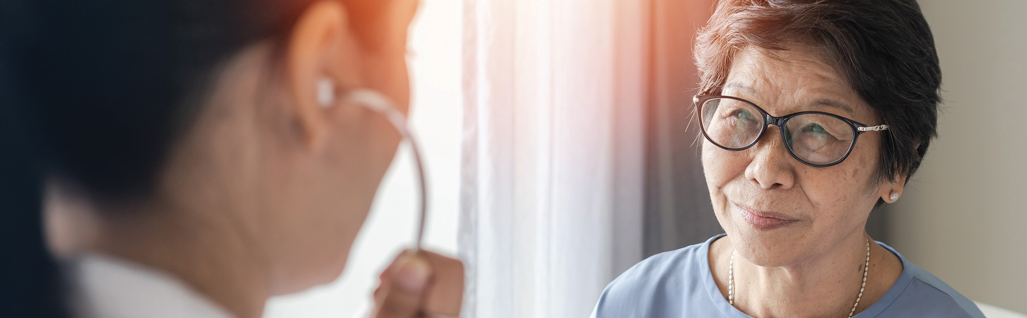 Caregiver using a stethoscope while speaking with an older woman, providing attentive, in-home medical support