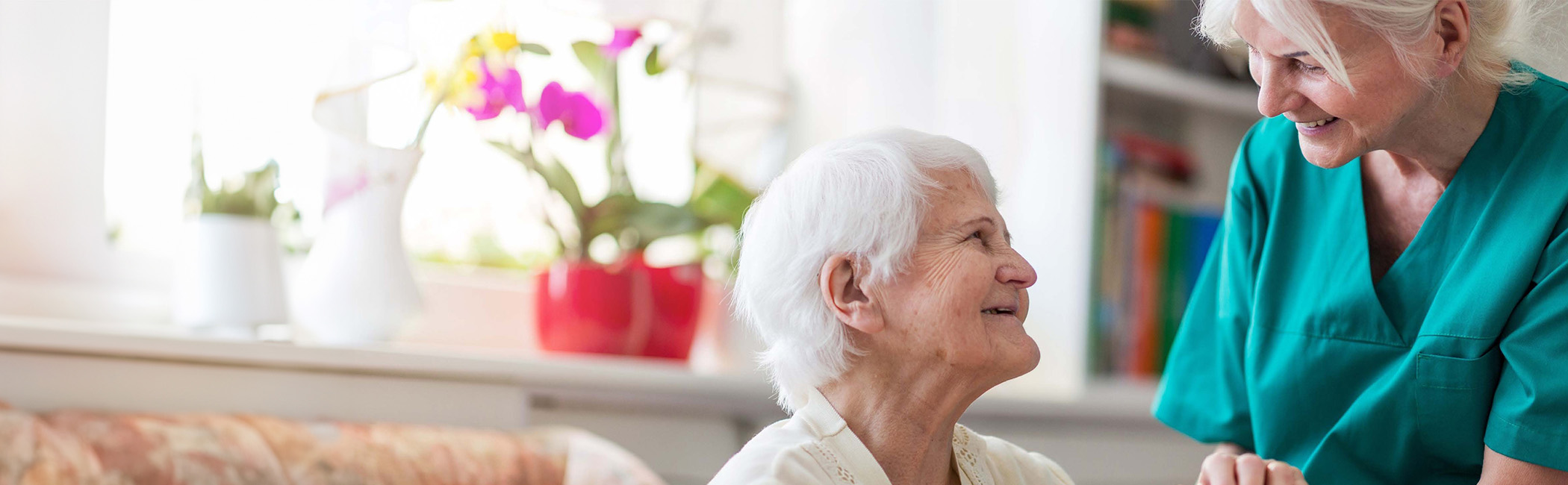 Caregiver leaning in and smiling as an older woman looks up at her, sharing a warm, reassuring moment in a bright home setting