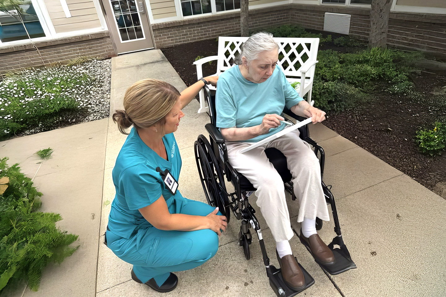 Caregiver kneeling beside a woman in a wheelchair outdoors, offering attentive support during a daily activity