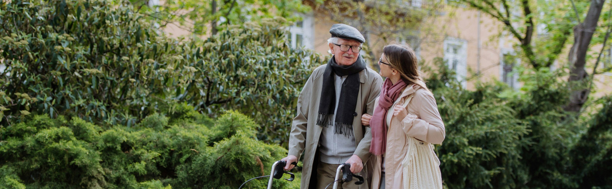 Older man using a walker while walking outdoors with a companion, supported with steady guidance and companionship