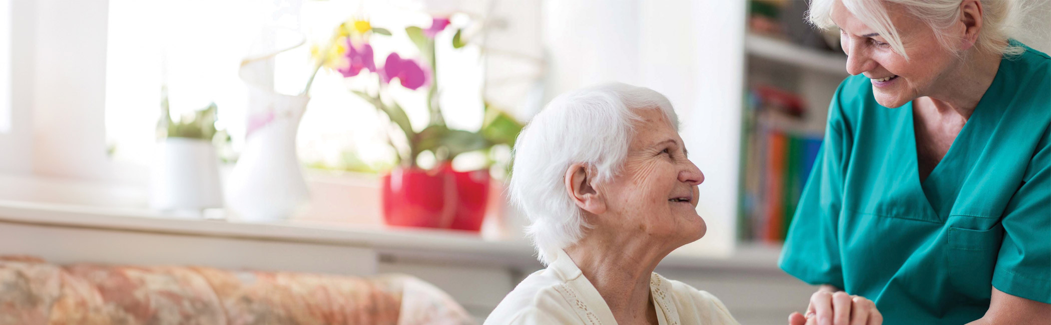 Caregiver leaning close to speak with an older woman, sharing a warm and attentive conversation in a comfortable home setting