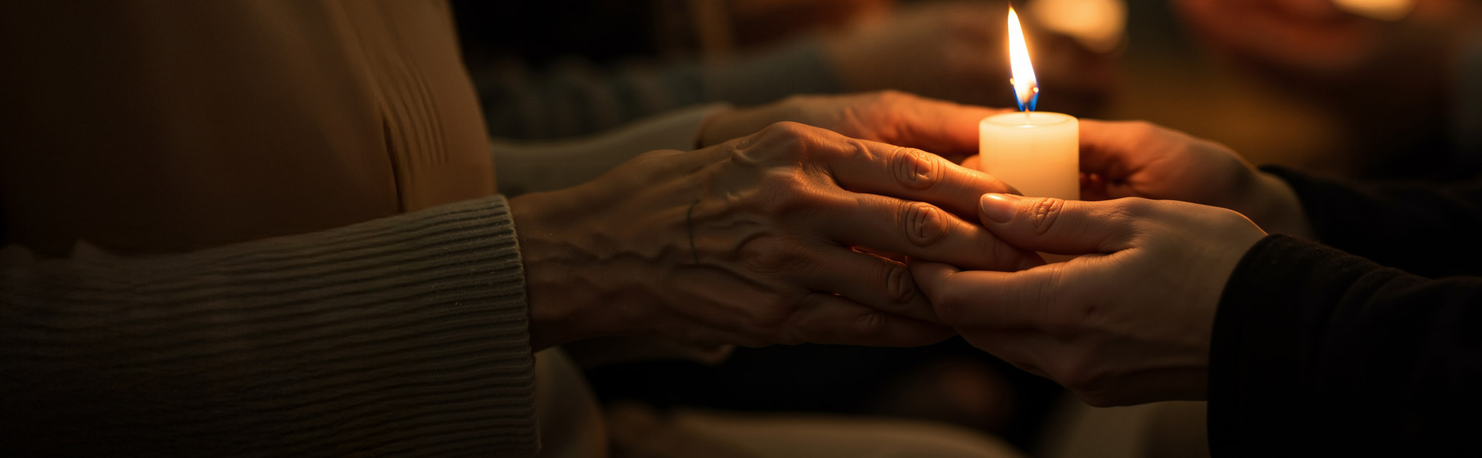 Hands passing a lit candle between two people, symbolizing remembrance, comfort, and shared presence