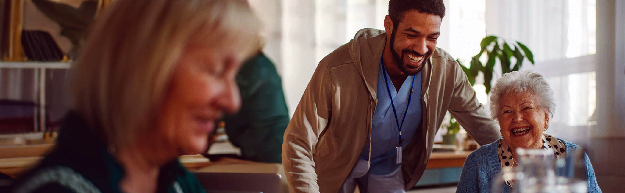 Caregiver leaning in with a smile as an older woman laughs at the table, sharing a joyful and uplifting moment