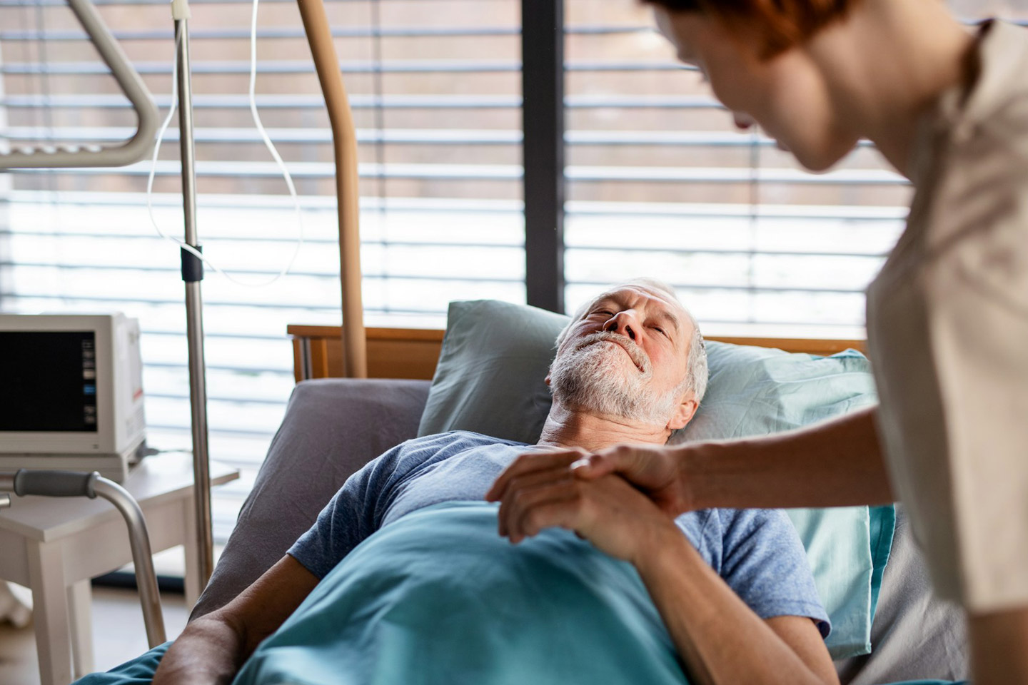 Caregiver holding an older man’s hand at the bedside, offering quiet comfort and presence during a moment of care