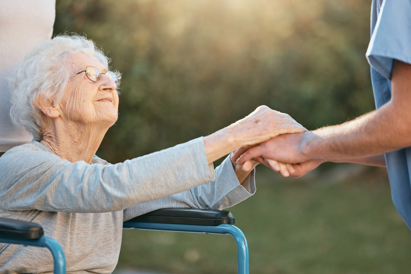 Caregiver gently holding an older woman’s hand outdoors as she sits in a wheelchair, sharing a moment of trust and reassurance