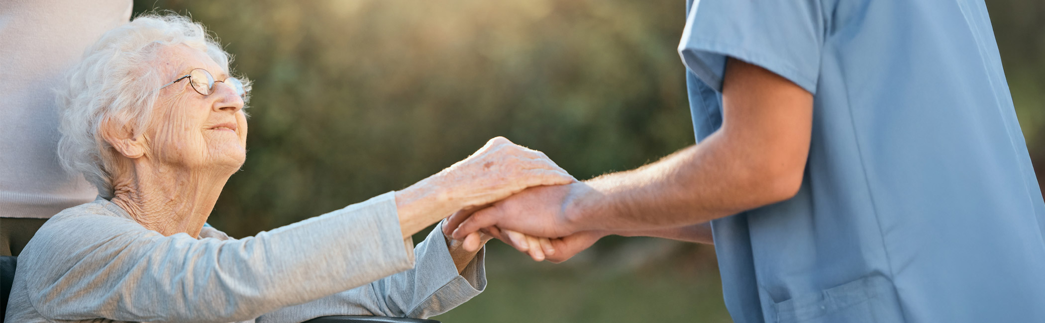 Caregiver gently holding an older woman’s hand outdoors as she sits in a wheelchair, sharing a moment of trust and reassurance