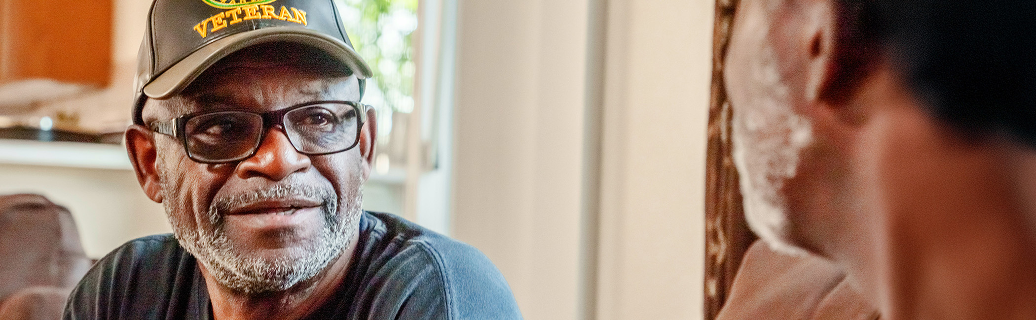 Older man wearing a veteran cap speaking with another person, sharing a warm, attentive conversation in a comfortable home setting