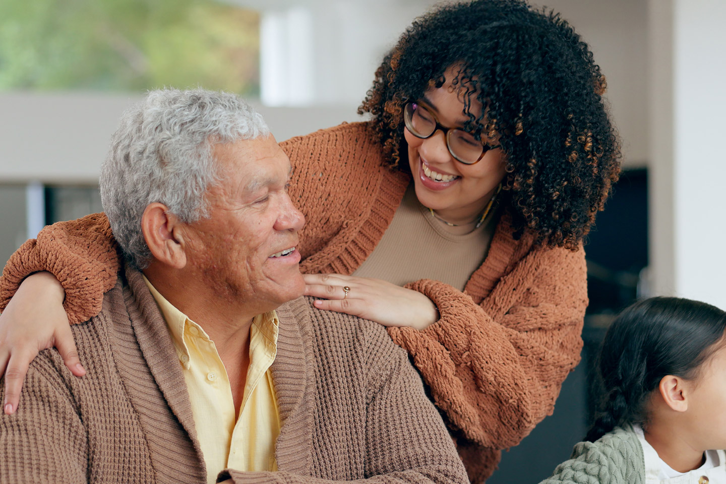 Family member leaning in with a smile and a hand on an older man’s shoulder, sharing a warm, supportive moment at home