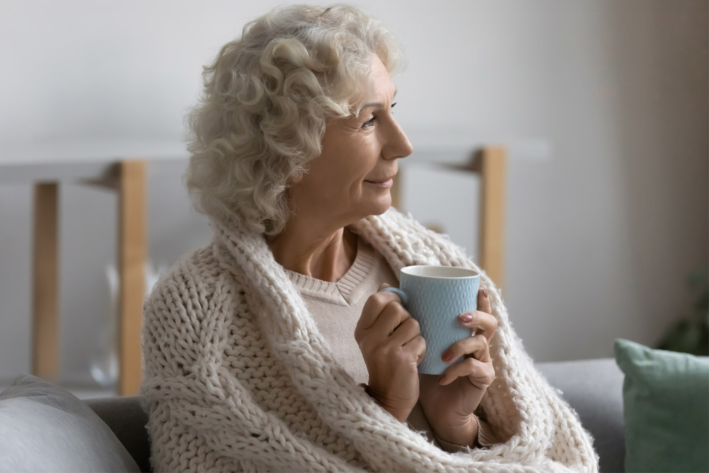 Older woman wrapped in a blanket holding a warm drink, sitting comfortably and enjoying a quiet, peaceful moment at home