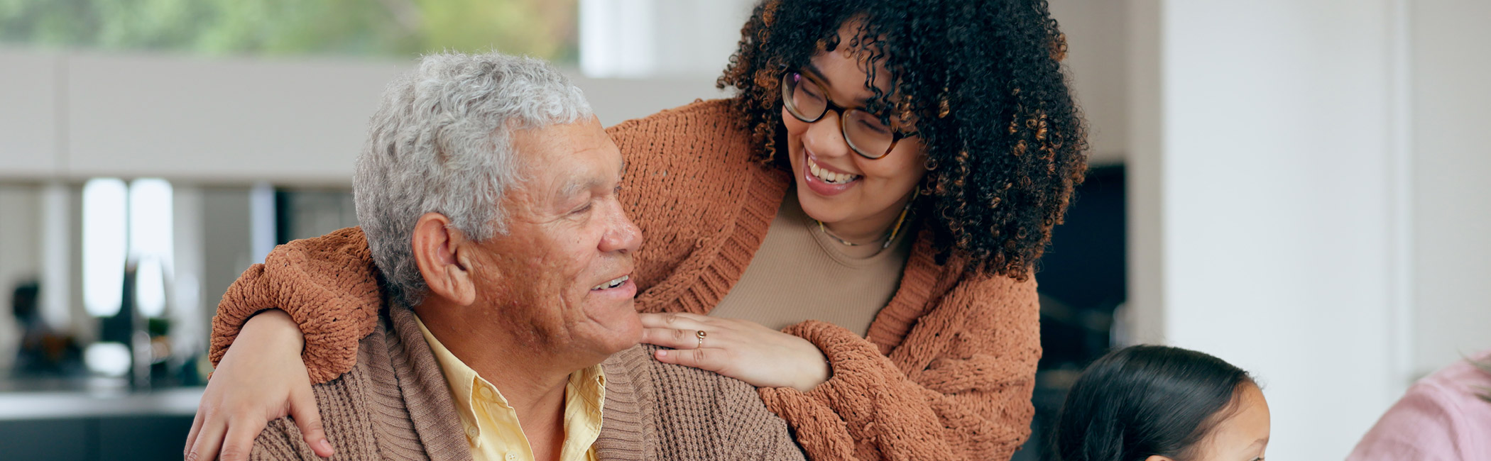Family member leaning in with a smile and a hand on an older man’s shoulder, sharing a warm, supportive moment at home
