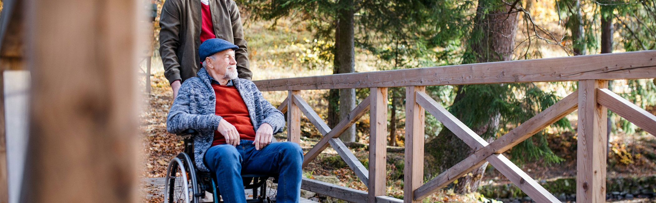 Caregiver pushing an older man in a wheelchair along a wooded path, supporting safe movement and time outdoors