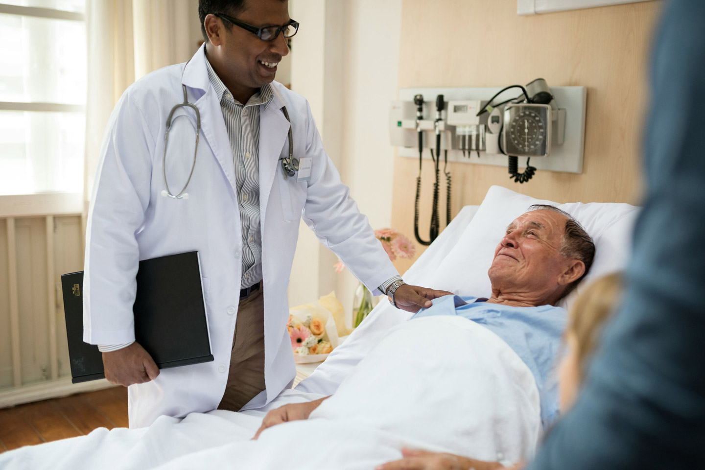 Doctor standing at the bedside with a hand on a patient’s shoulder, offering reassurance and compassionate medical care
