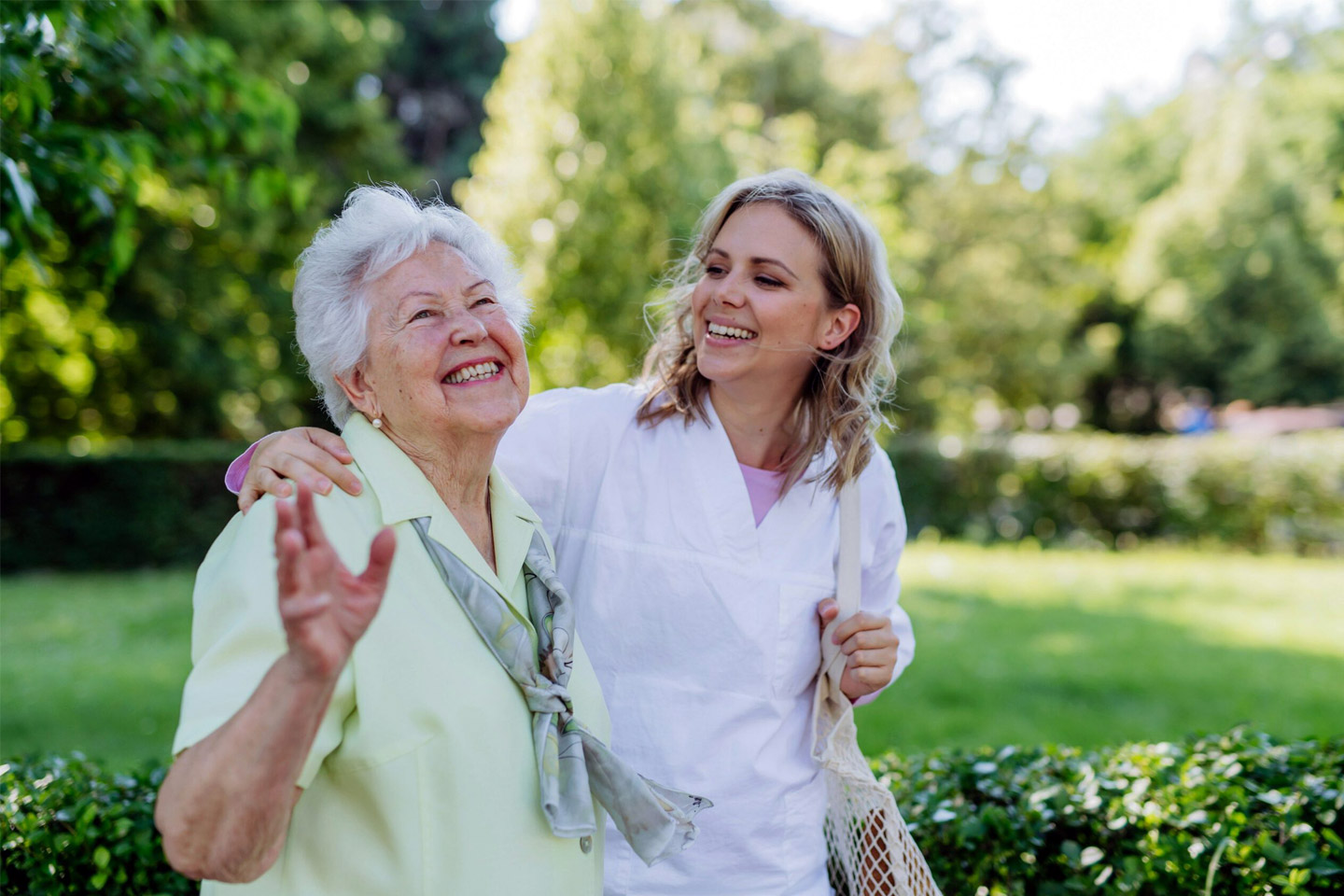 Caregiver walking beside an older woman outdoors, sharing a joyful moment and supportive companionship