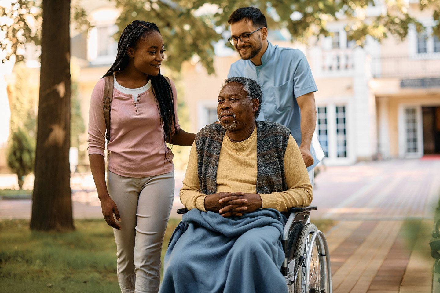 Caregiver and companion walking alongside a man in a wheelchair outdoors, providing steady support and companionship