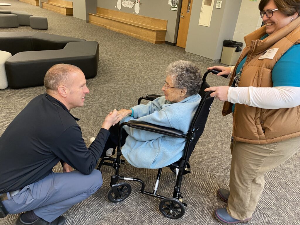 Millie Ferro, a former Concord High School coach and teacher, smiles while seated in a wheelchair and shaking hands with a man during a heartfelt return visit, accompanied by a Luminary Hospice care nurse.